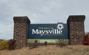 A road-sign sign reads, "Welcome to Maysville: Naturally Welcoming" 