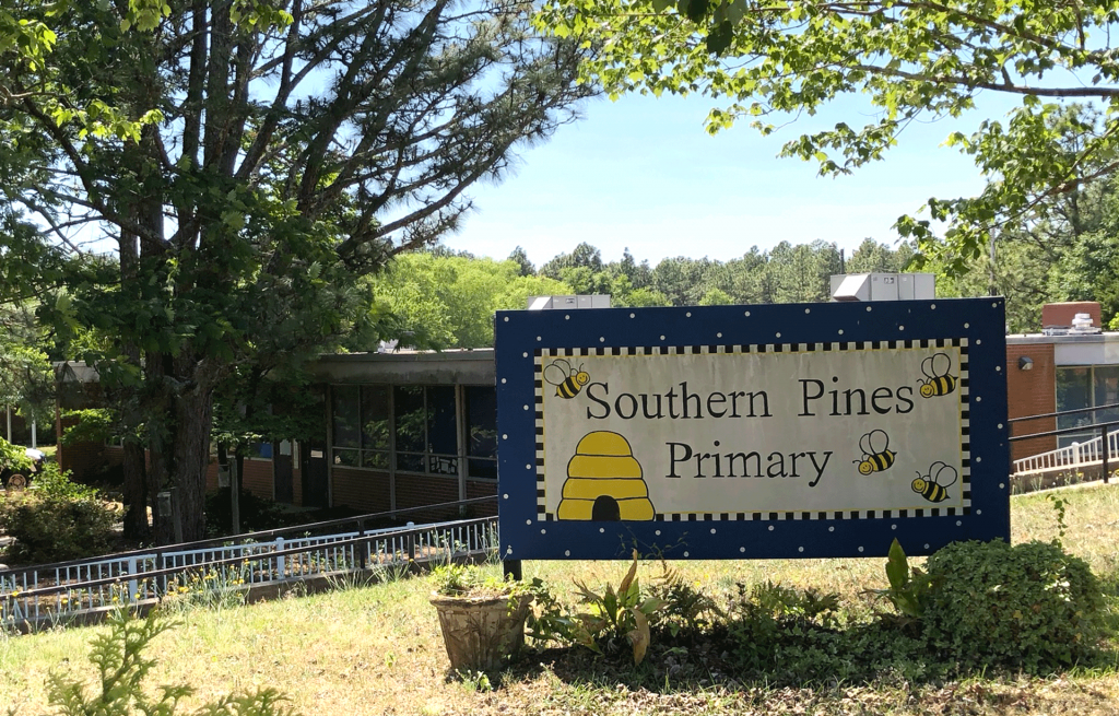 A painted sign in front of a brick building reads "Southern Pines Primary" with cartoon bumblebees and a blue border.